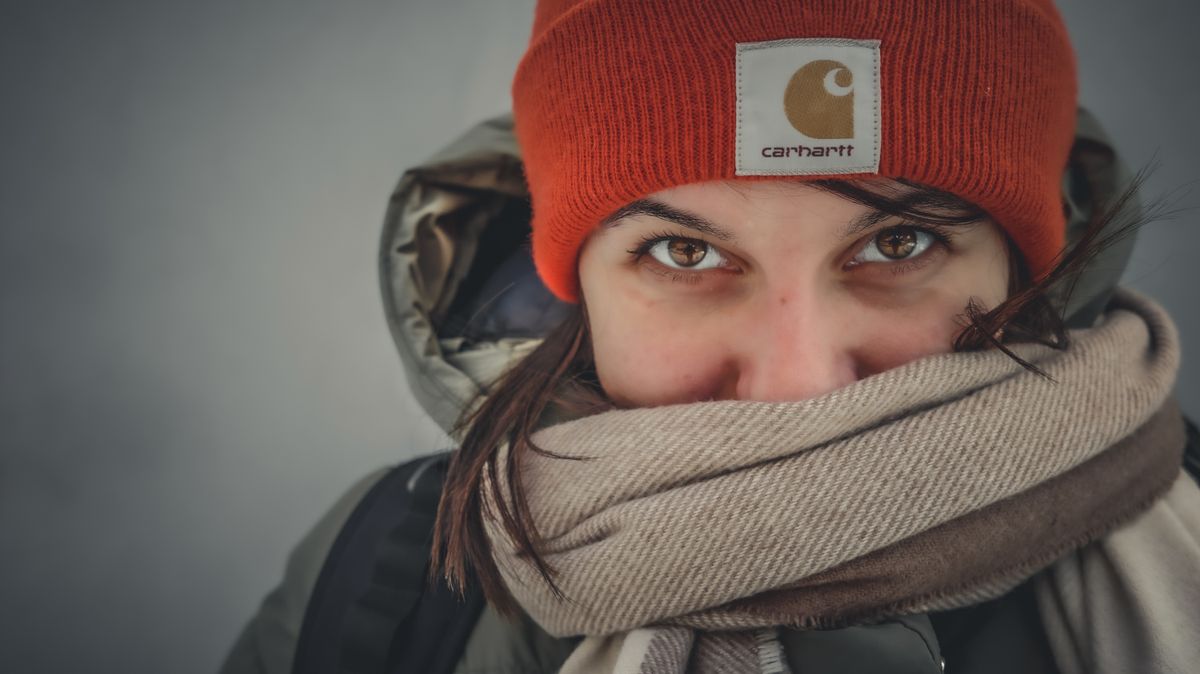 Na zdjęciu stoi osoba w pomarańczowej zimowej czapce, owinięta szalikiem, tak, że widać tylko oczy.
